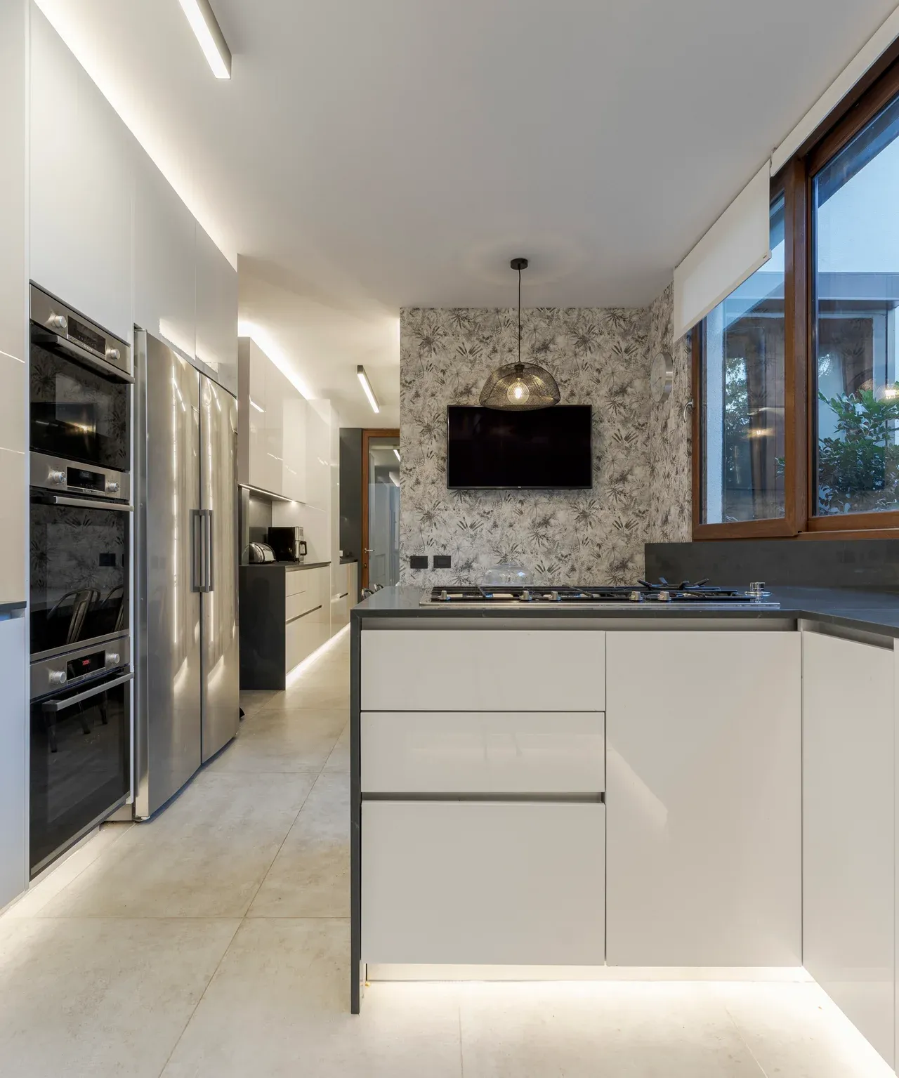 Diseño de cocina moderna con gabinetes blancos minimalistas y sin tiradores. Incluye una isla con encimera oscura y placa de cocción, y una columna con horno doble y refrigerador de acero inoxidable. El fondo presenta una pared con papel tapiz gris monocromático y una pequeña televisión, y una gran ventana de madera a la derecha