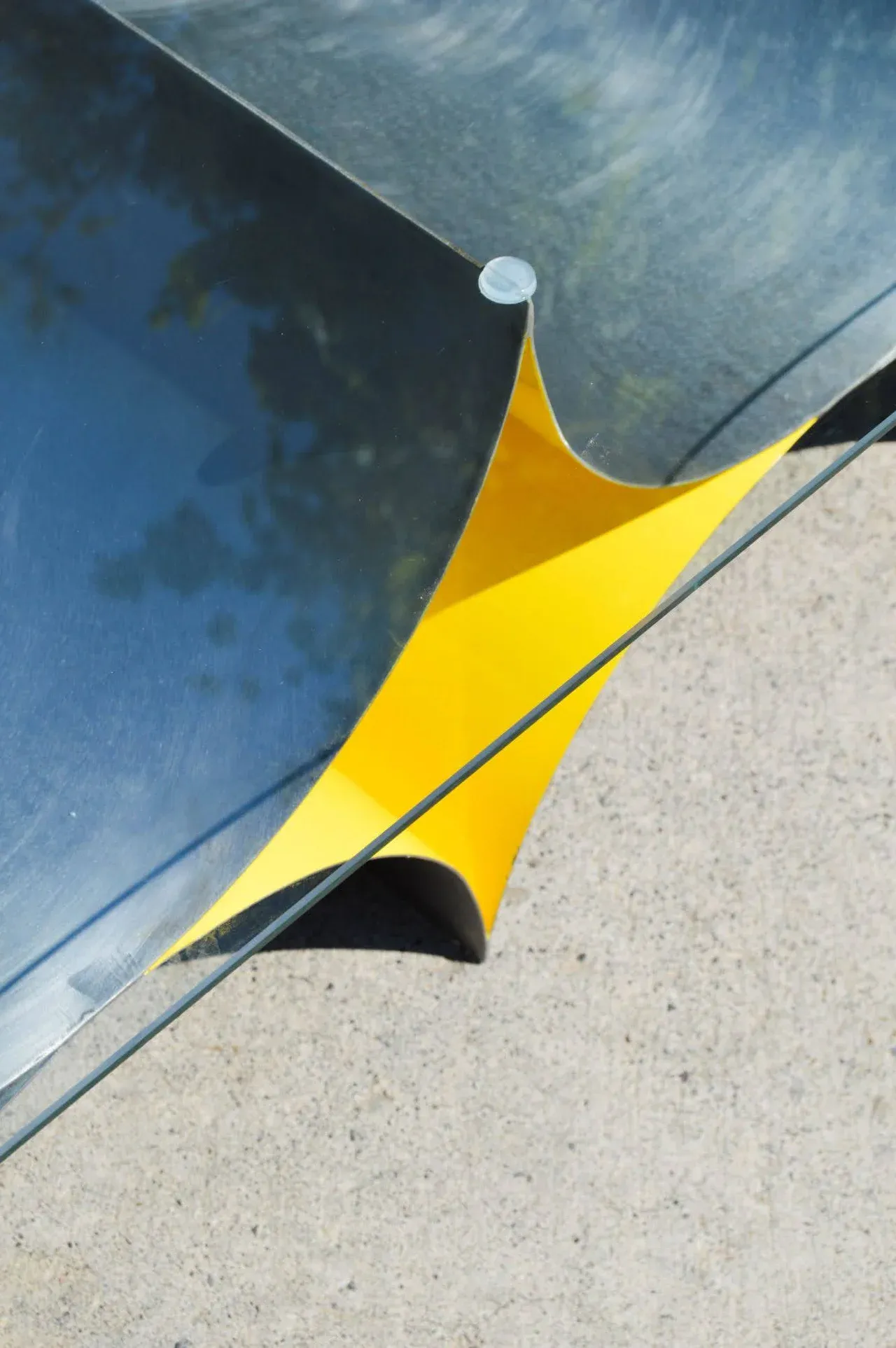 Primer plano de un detalle constructivo de la Mesa 00. Se muestra la unión del acero bruñido, el acento interior electropintado en amarillo brillante, y la tapa de vidrio templado. El diseño resalta las líneas limpias y los materiales industriales de esta pieza de mobiliario minimalista