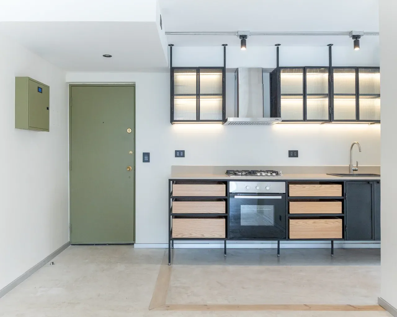 Detalle de cocina minimalista en Las Condes con puerta verde oliva, gabinetes industriales con cajones de madera y vitrinas colgantes de vidrio estriado