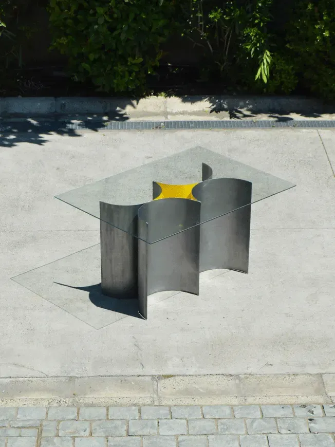 Mesa de centro con base escultórica de acero curvado y tapa cuadrada de vidrio, destacando el diseño de mobiliario moderno