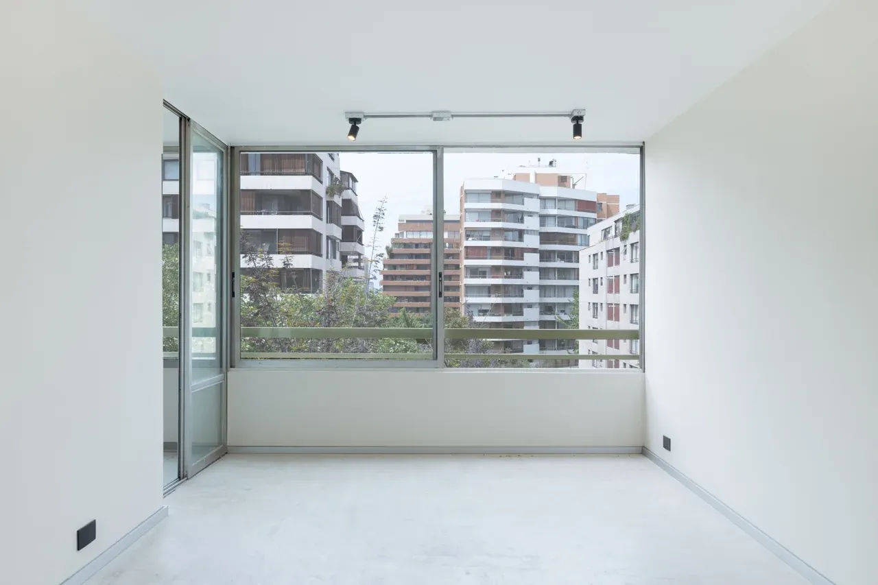 Interior de departamento remodelado en Las Condes. Espacio vacío, blanco y minimalista con piso de porcelanato claro y ventanales de piso a techo que ofrecen vista a edificios residenciales. El diseño maximiza la luz natural y presenta una estética sobria y contemporánea, destacando la luminosidad del ambiente