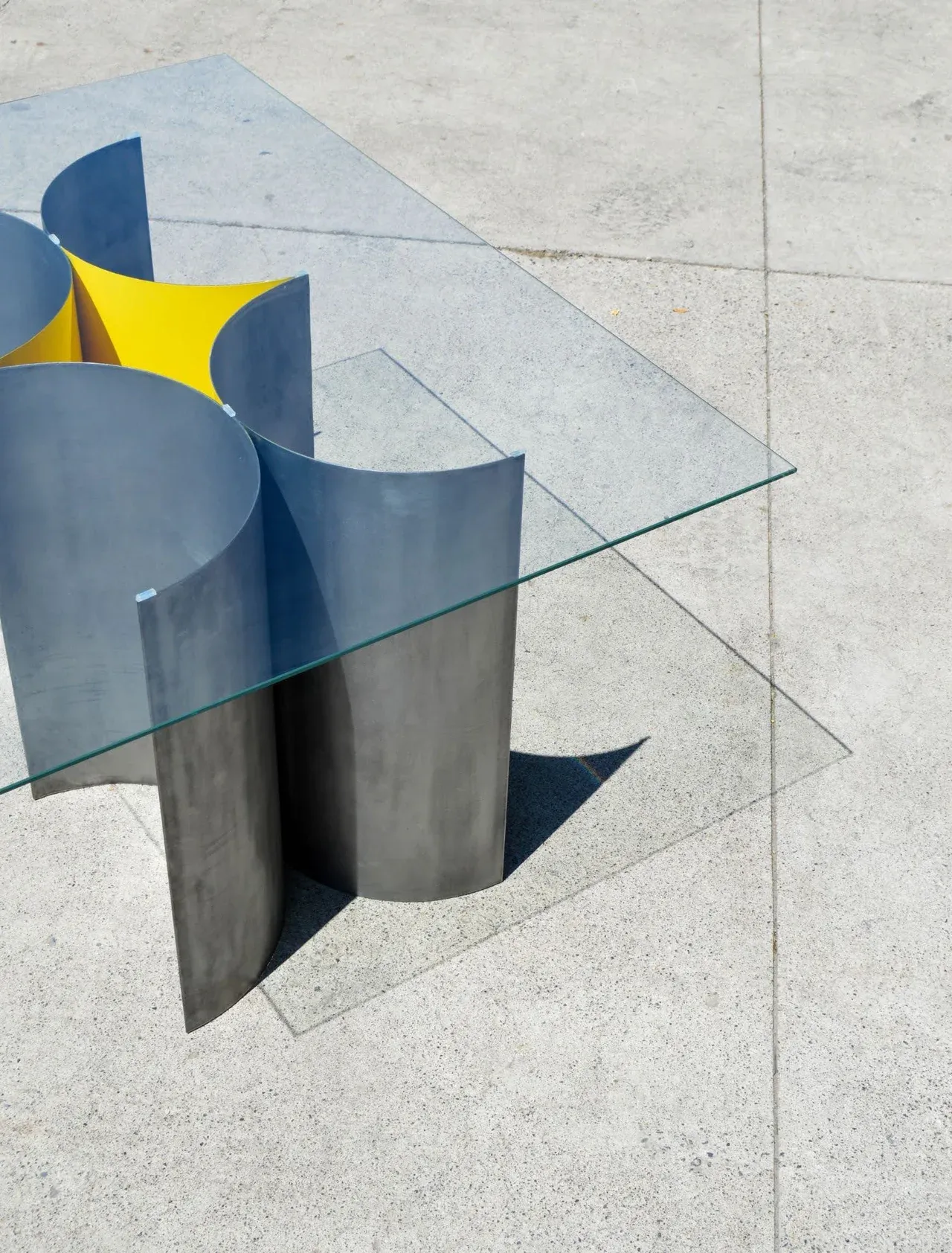 Detalle de la Mesa 00, un diseño escultórico y minimalista. La base está compuesta por paneles curvos de acero bruñido gris, con acentos internos en amarillo vibrante, y cubierta por vidrio templado. Fotografía de la mesa sobre suelo de concreto claro, ideal para mobiliario contemporáneo