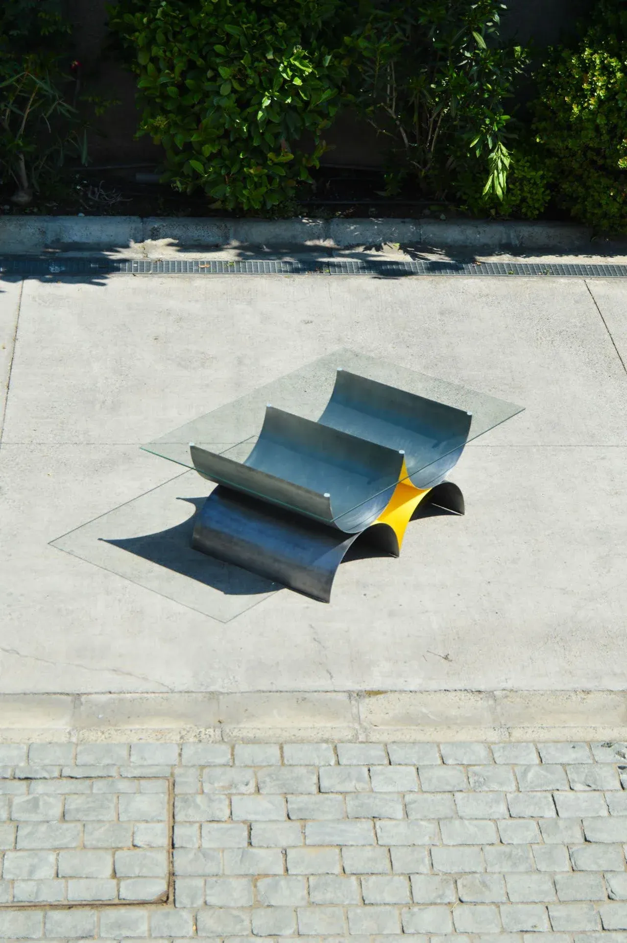 Vista aérea de la Mesa 00, una mesa de centro de diseño escultórico con base de acero bruñido curvado y acento amarillo. La cubierta es de vidrio rectangular. La mesa se exhibe sobre un pavimento de concreto claro, con una franja de adoquines de ladrillo en primer plano, destacando su versatilidad para exteriores o interiores