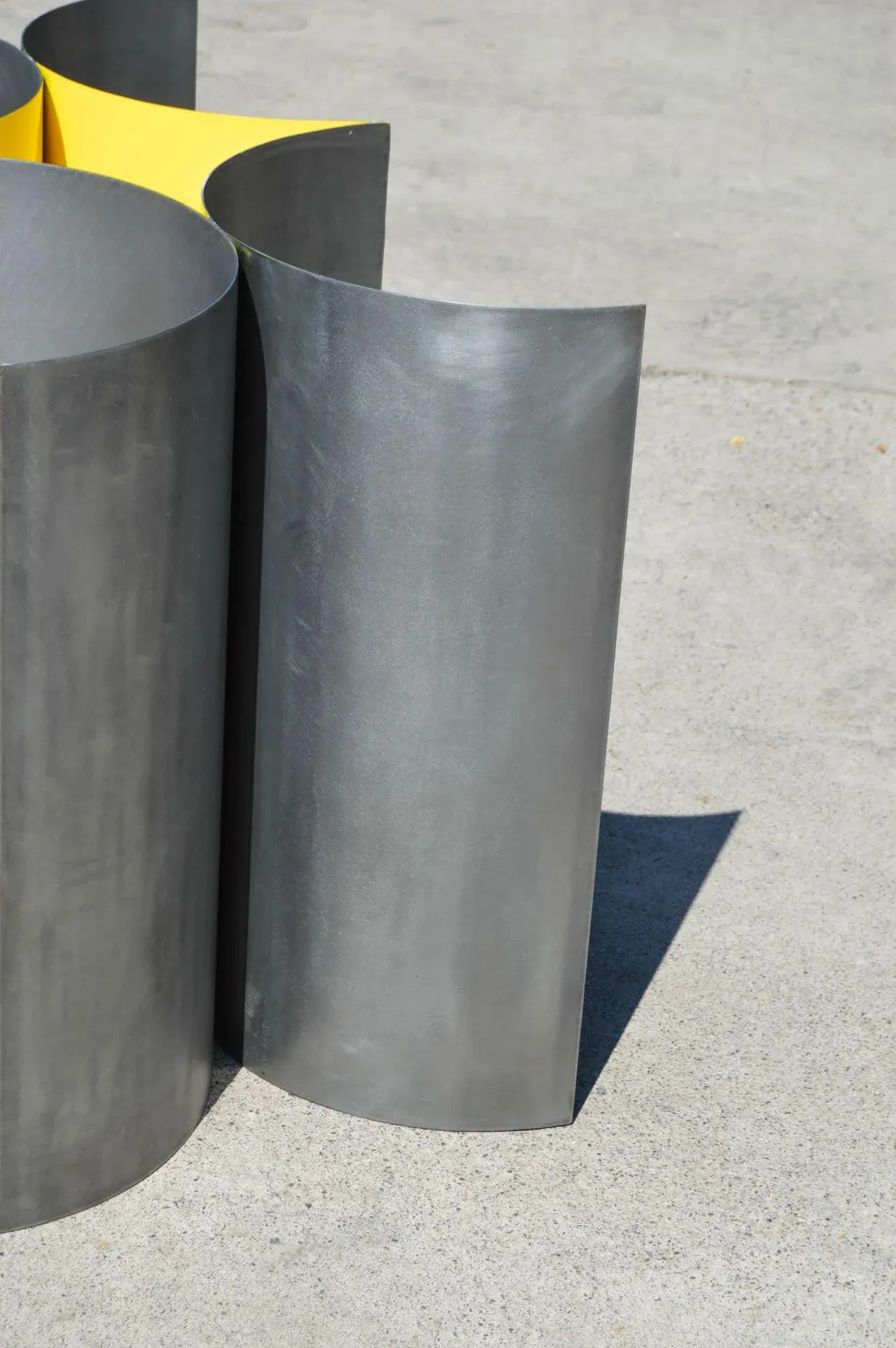 Primer plano del detalle de la base de la Mesa 00, destacando la textura y acabado del acero bruñido curvado. La luz del sol incide sobre el metal gris oscuro, proyectando una sombra nítida sobre el suelo de concreto. Se aprecian detalles del diseño minimalista y la ingeniería de la base escultórica